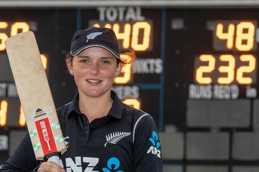 A picture of NZ cricketer Amelia Kerr with bat over shoulder and scoreboard behind
