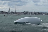 The hull of a yacht lays on the surface of the water after it topples over, as another small yacht approaches in the distance