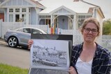 A woman holding a black and white drawing of a house, standing in front of the same real house.