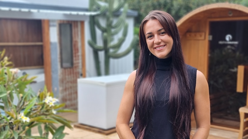 A young woman with long dark brown hair and a black shirt smiling and standing in her backyard