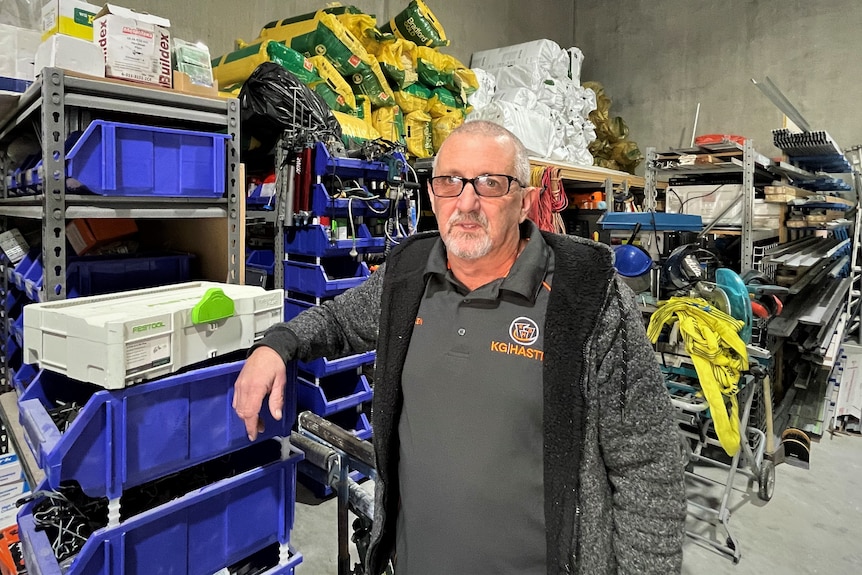 A man stands in his shed with plastering supplies on shelves and the floor behind him 