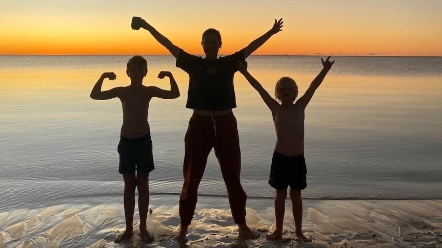 A woman and two kids stand beside the shoreline as a sunset lowers behind them.