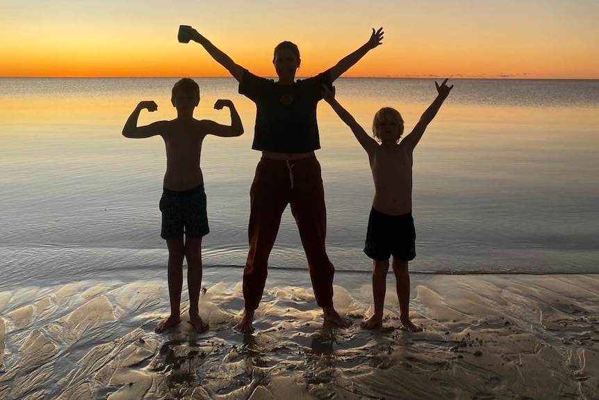 A woman and two kids stand beside the shoreline as a sunset lowers behind them.