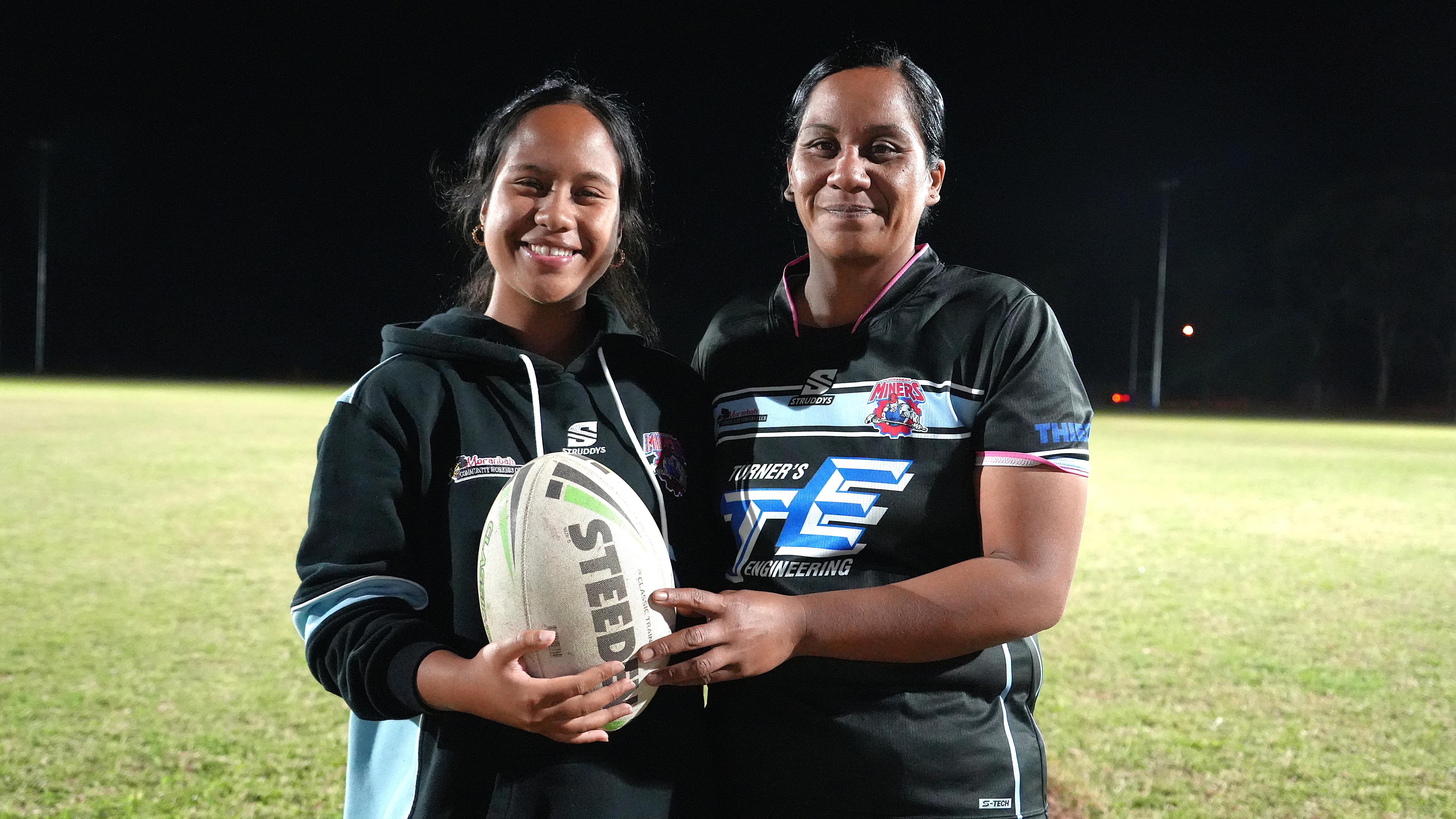 Mother and daughter with football smiling at camera, in uniform, on footy field at night time 