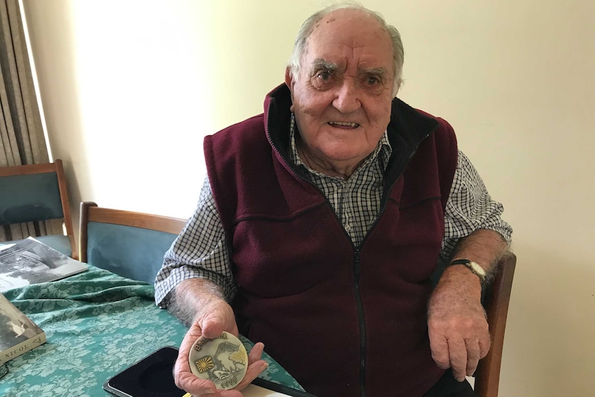 Elderly Max Lugsdin sits in a chair holding a silver medallion engraved with Mt Fuji, a Japanese war flag and the year 1966.