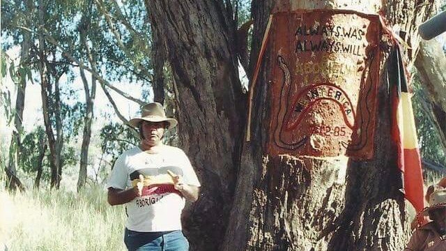 William Bates stands in front of a tree with "always was always will be Aboriginal land" carved in to it.