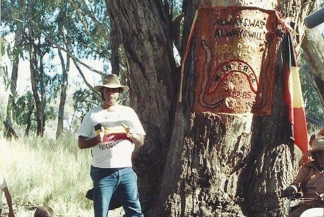 William Bates stands in front of a tree with "always was always will be Aboriginal land" carved in to it.