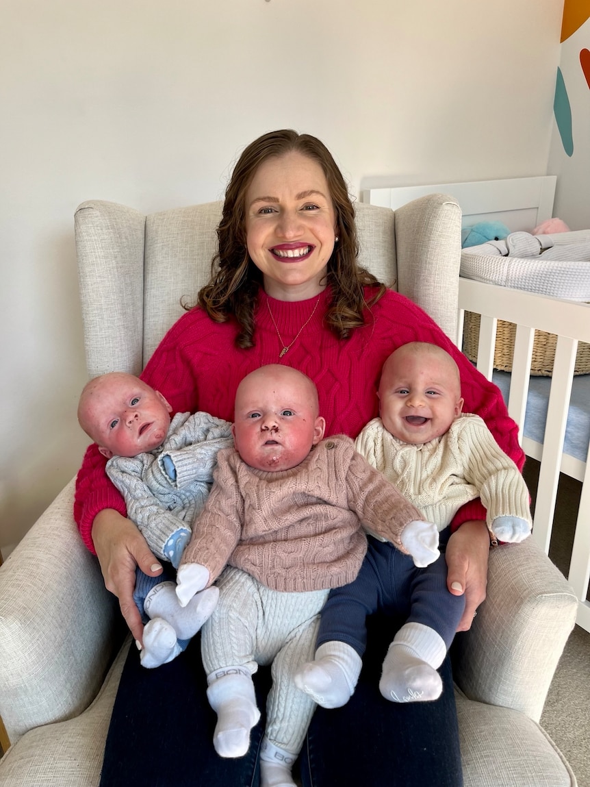 A woman sits in a chir smiling with her three babies on her lap.