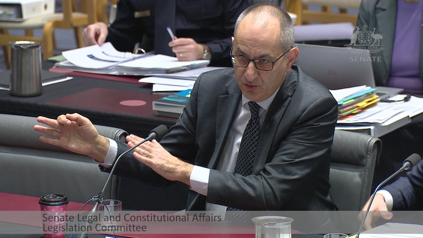 Mike gestures, standing in parliament wearing a suit and tie.