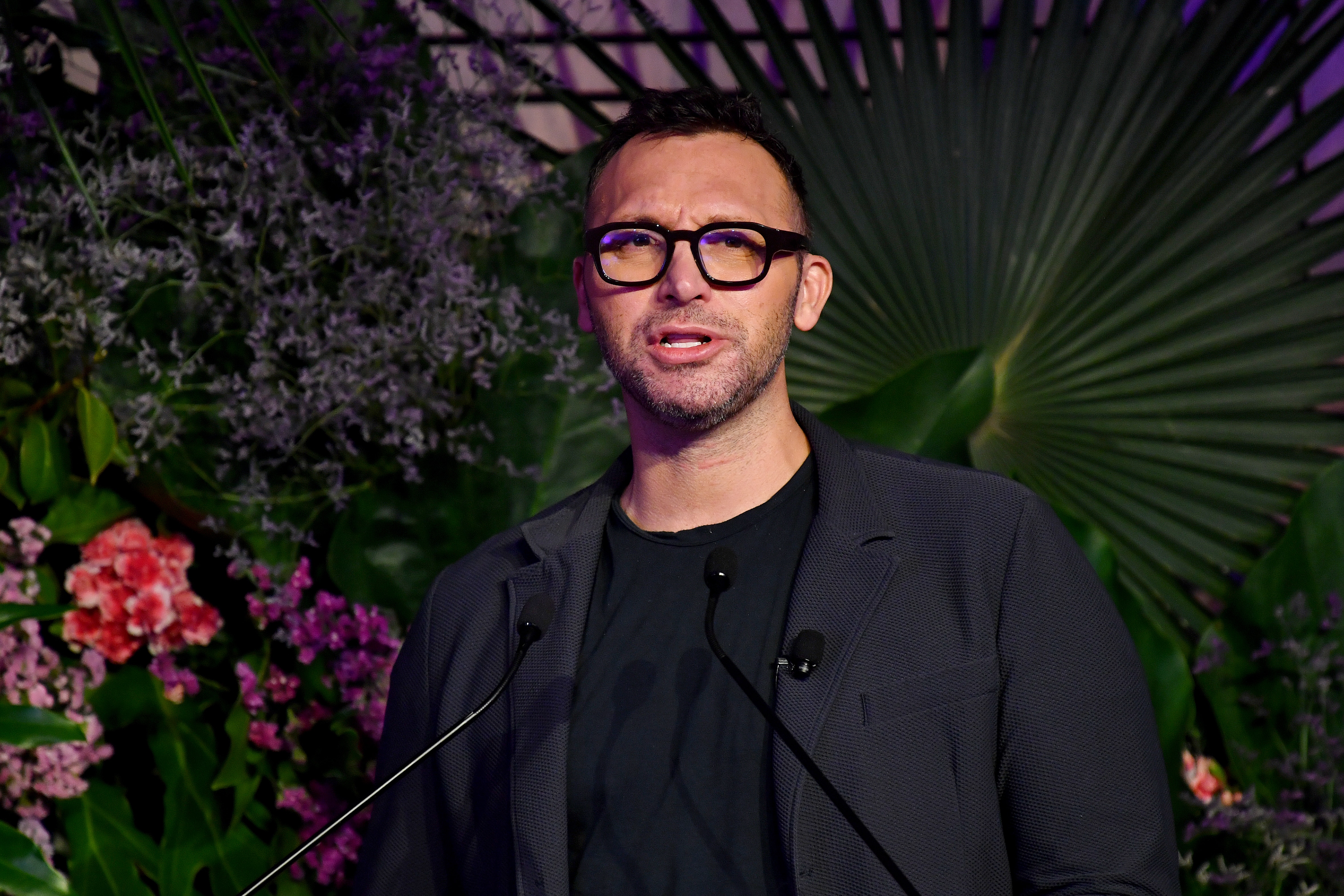 Ian Thorpe in a suit and glasses talking to media with a serious expression in front of a wall of flowers.