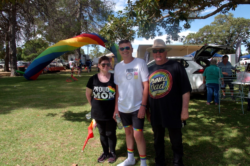 A man in a shirt and shorts stands between his mum and dad in front of a blow-up rainbow at a park.