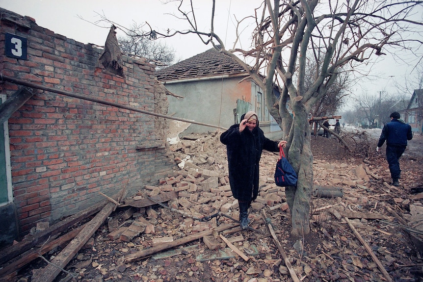 A woman wearing a black coat and head scarf walks over the rubble of a destroyed building. 