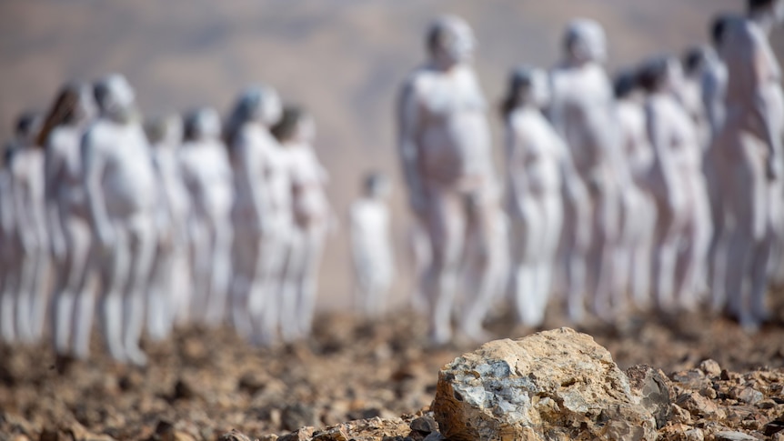 A rock sits in the foreground in the desert as naked people painted white stand, blurry, in the background.