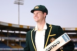 Steve Smith stands and smiles in a blazer holding a cricket bat