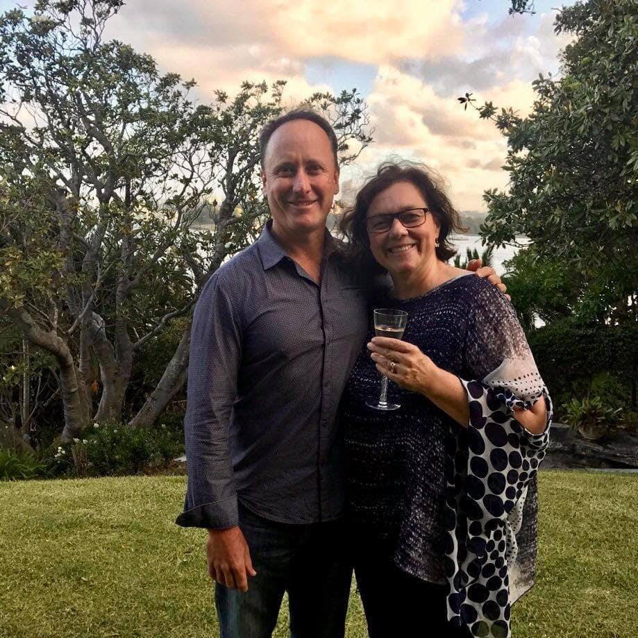 Tim stands with his arm around Lynelle in a garden, she holds a glass of wine. Water can be seen in the distance behind them.