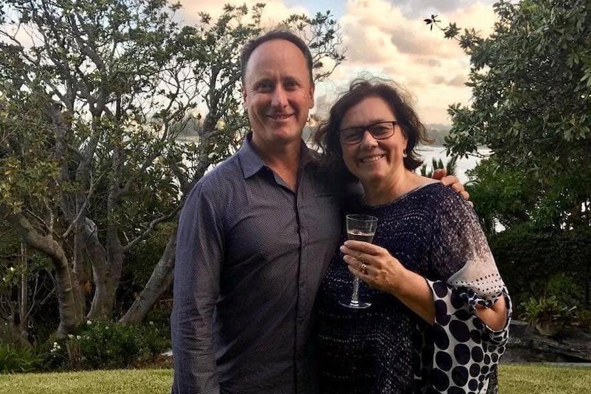 Tim stands with his arm around Lynelle in a garden, she holds a glass of wine. Water can be seen in the distance behind them.