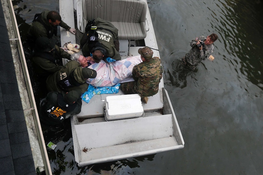 An elderly woman is seen from above being helped in a boat by six uniformed men.