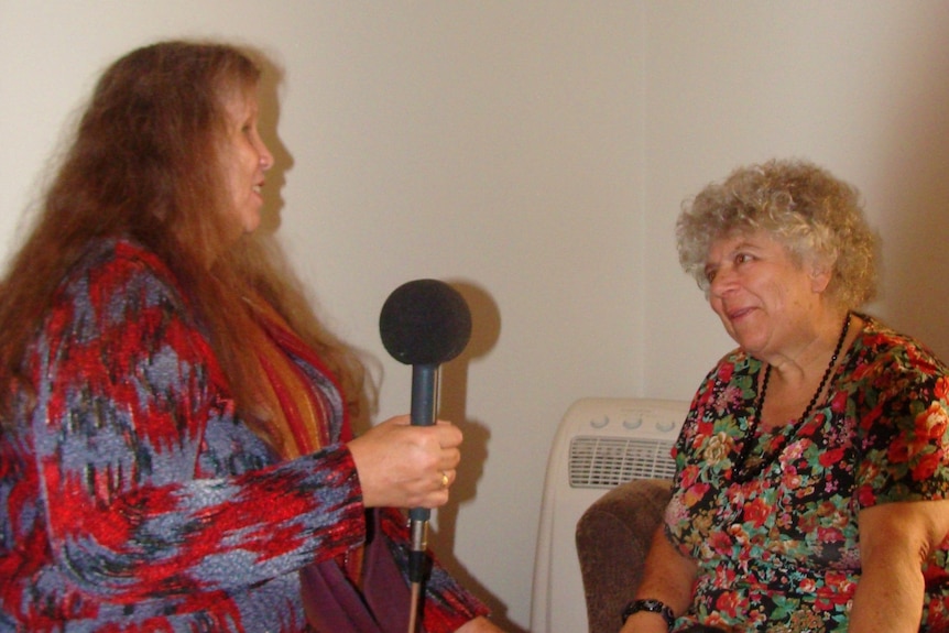 Elaine sits across from Miriam with a microphone in her hand. Miriam has one hand on the black guide dog while looking at Elaine