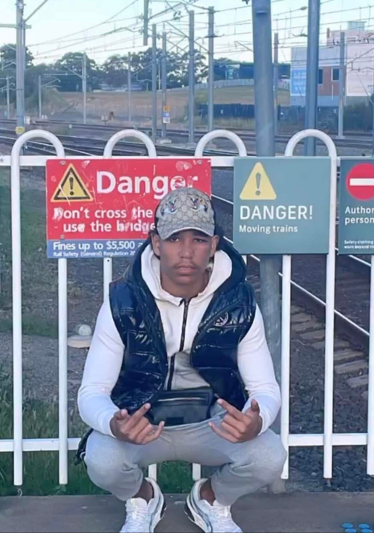 Young man bent down with findergs up at train station platform with danger signs behind him