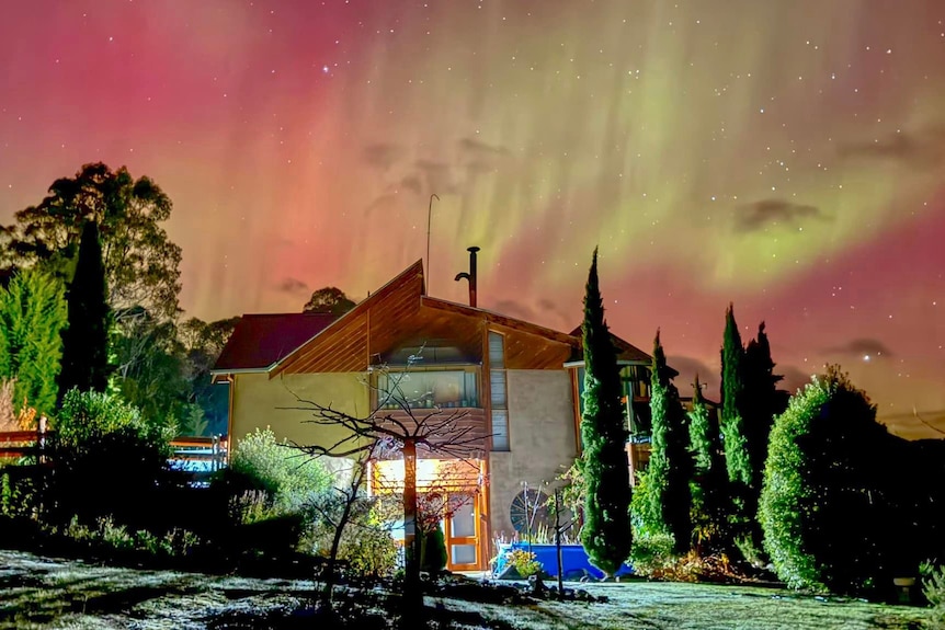 An orange and green aurora is seen behind a two-storey house that is illuminated by a flash