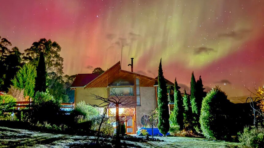 An orange and green aurora is seen behind a two-storey house that is illuminated by a flash