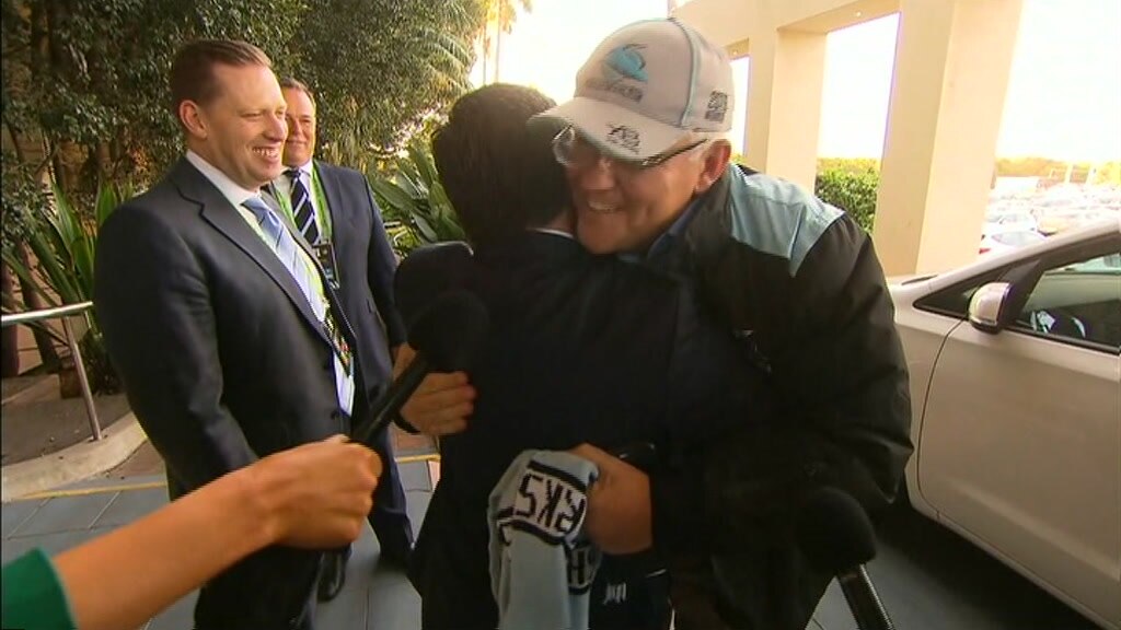 Scott Briggs in a suit and tie on the left watches and smiles as Scott Morrison gives another man a hug 