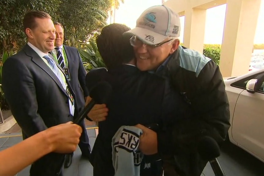 Scott Briggs in a suit and tie on the left watches and smiles as Scott Morrison gives another man a hug 