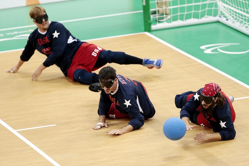 Members of the US goalball team lie on the court wearing blackout goggles trying to block the ball from going into the net.