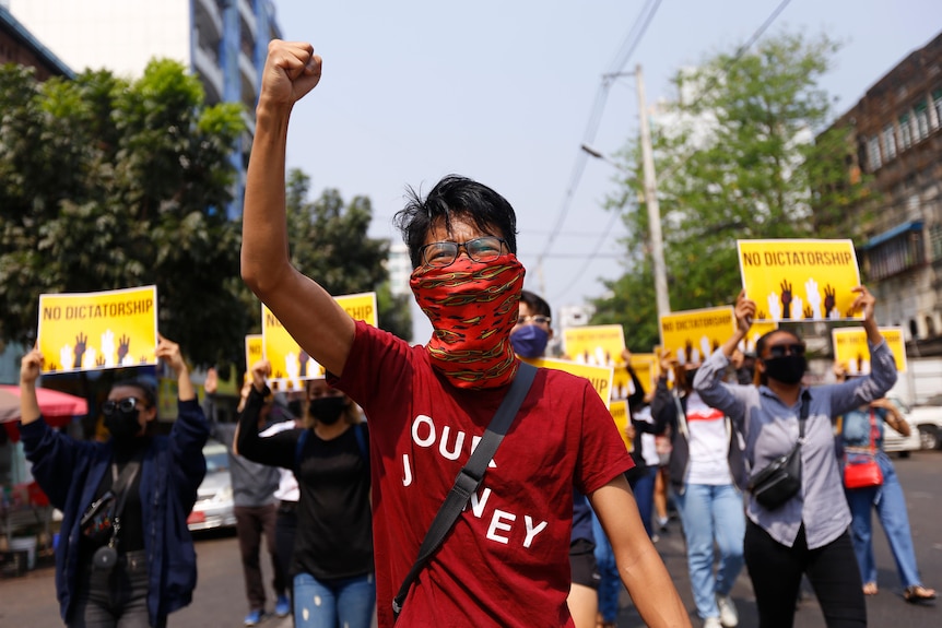 An anti-coup protester holds up his fist as a crowd holding signs marches behind him.