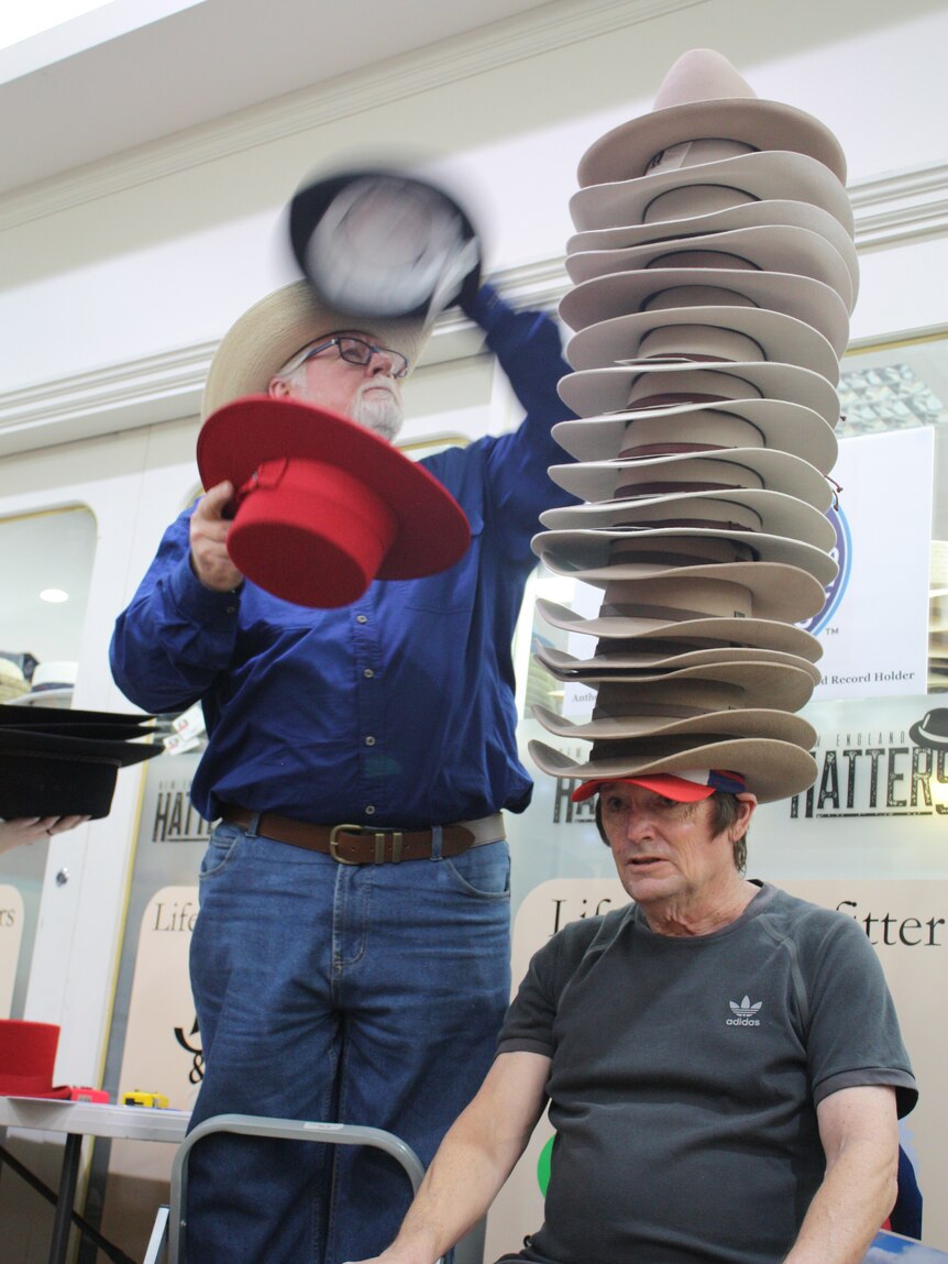 A man sits on a chair with a large stack of hats on his head, as someone brings another red hat towards the stack
