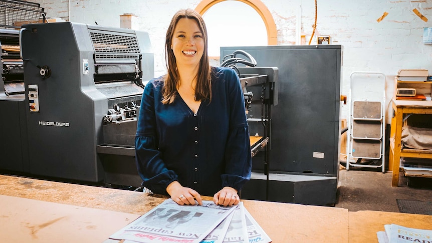 A woman in a dark blue top with long brown hair stands at a table with newspapers on it, with a printing press behind her.
