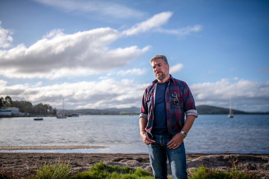 A man in a plaid shirt stands on the shore of a beach with the ocean and clouds in the background.