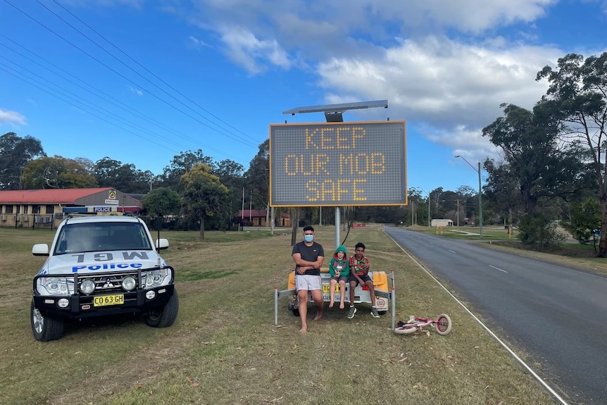 Aboriginal man and children sitting near a digital sign which reads "Keep our mob safe"