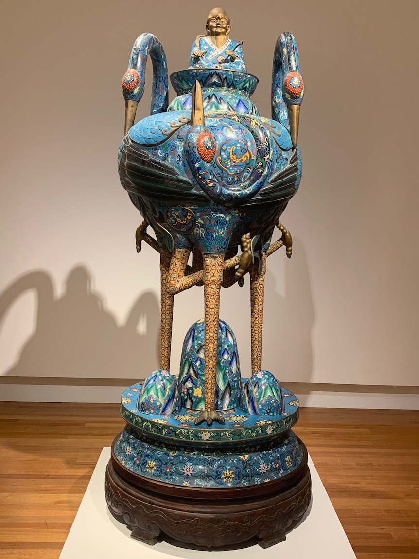 A blue and embellished urn, taller than an adult human, supported by three standing cranes (birds) on a wooden base.