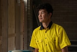 A man in a yellow work shirt looks out a window.