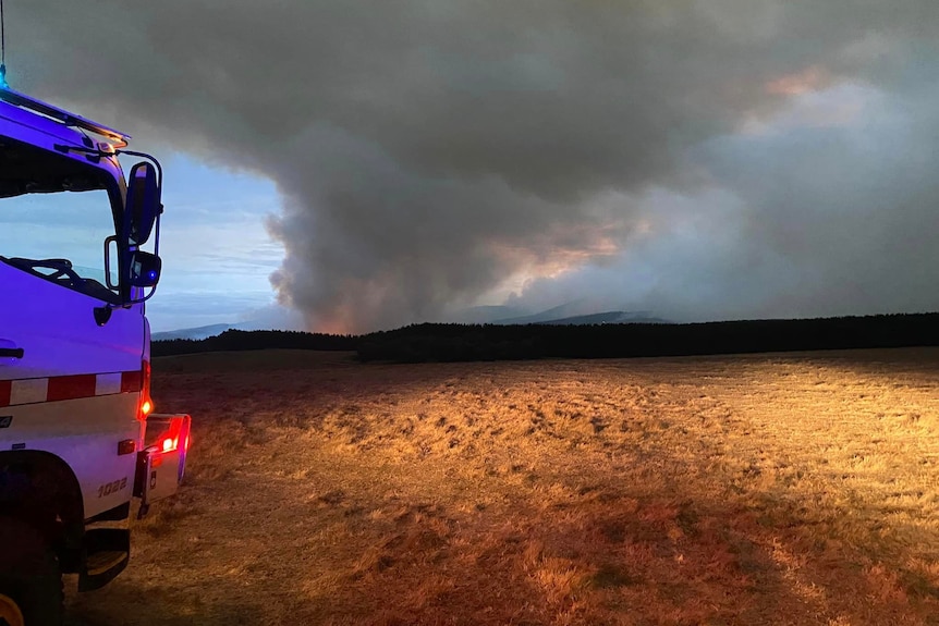 A fire truck in a field. In the distance a bushfire burns.