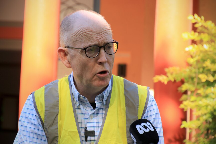 A middle-aged man wears a blue business shirt, yellow high-vis vest and black glasses.