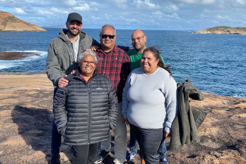 Three men and two women stand on a rocky shore near the ocean.