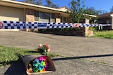 A low-set brick house with flowers outside, police tape cordons off the yard.