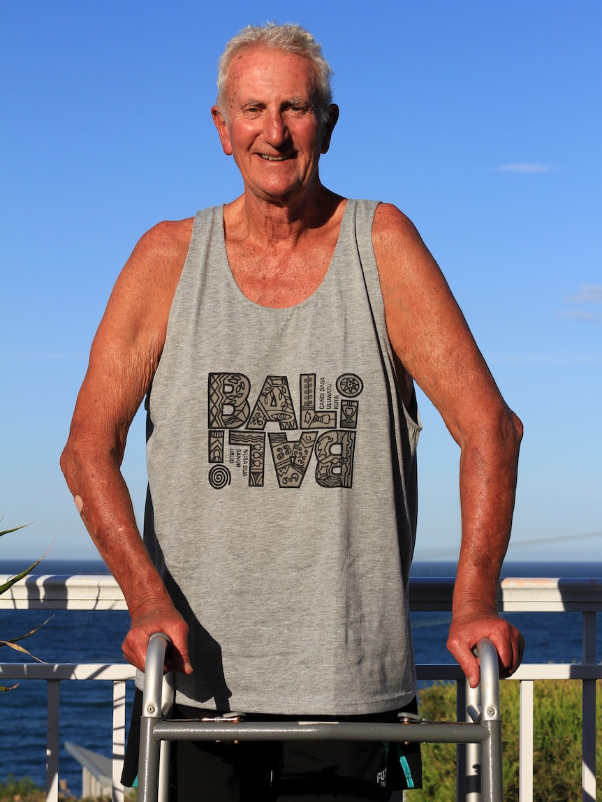 An elderly man holds a walker and stands on a balcony in front of the ocean.