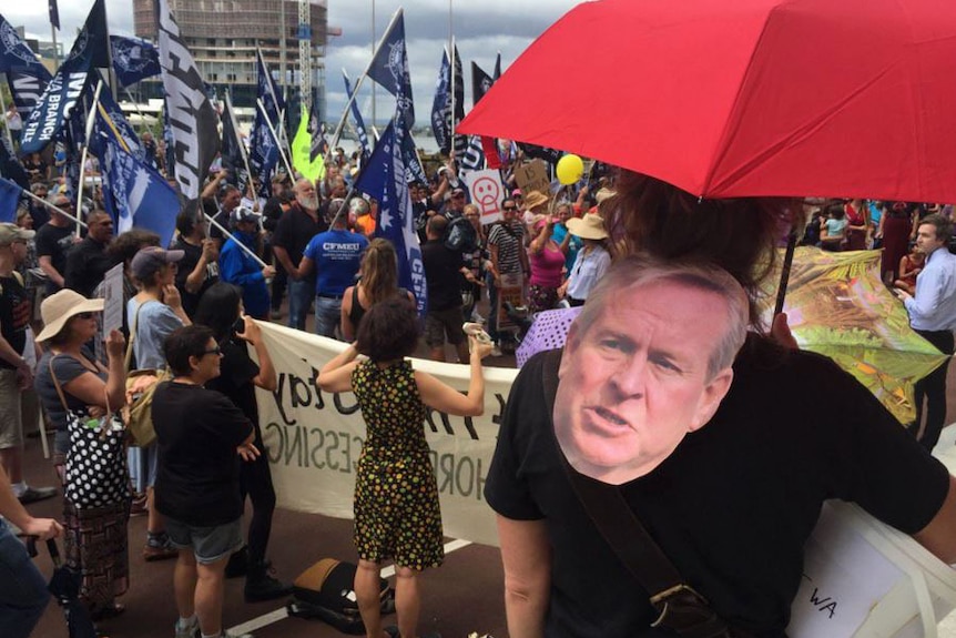 Crowds wave union flags and a man wears a mask of Colin Barnett over his shoulder at a rally outside Parliament House.
