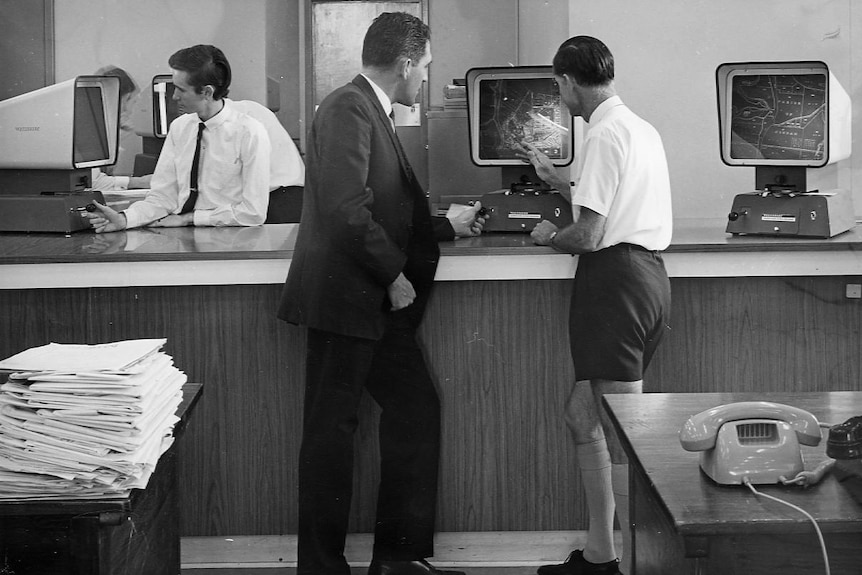 Three men in the 1960s at a desk working on microfilm.