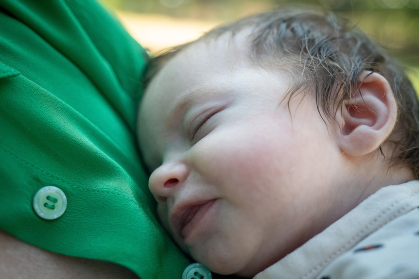 Close up of young sleeping baby with dark hair being held close to a green buttoned shirt.