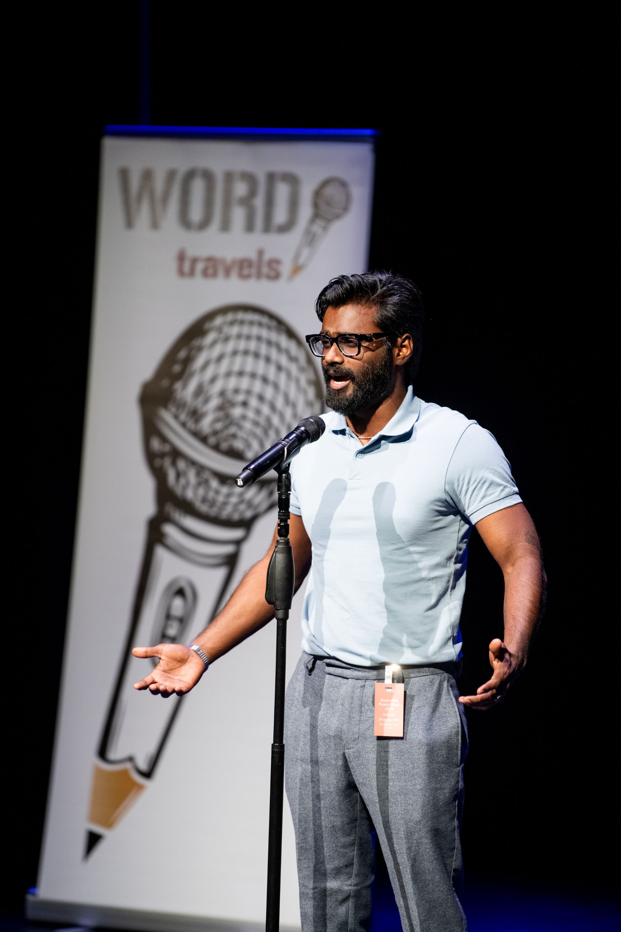 A man with dark hair, a beard and glasses, wearing a light-blue polo t-shirt, speaking into a microphone on stage.