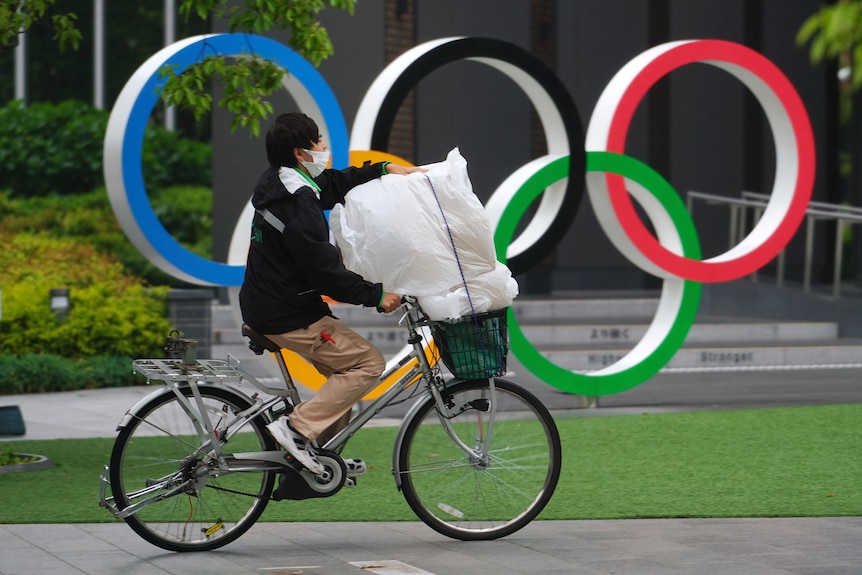  A man wearing protective masks rides a bicycle in front of he Olympic Rings in Tokyo.