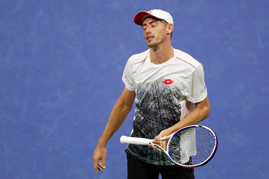 John Millman closes his eyes, looks disappointed, as he holds his racket loosely, standing on the blue court
