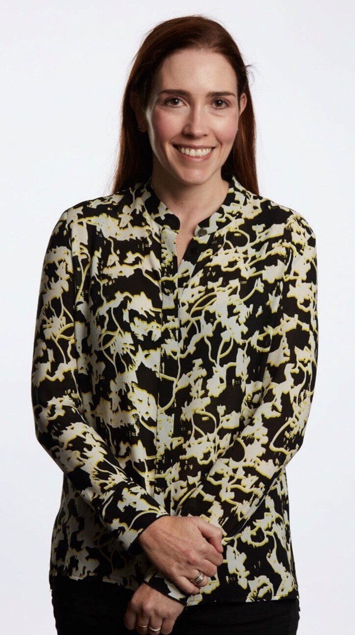A woman standing up and smiling with hands infront of each other. Wearing a black and green shirt
