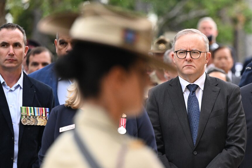 Mr Albanese looks serious, standing near soldiers and others in suit and tie at the outdoor ceremony.