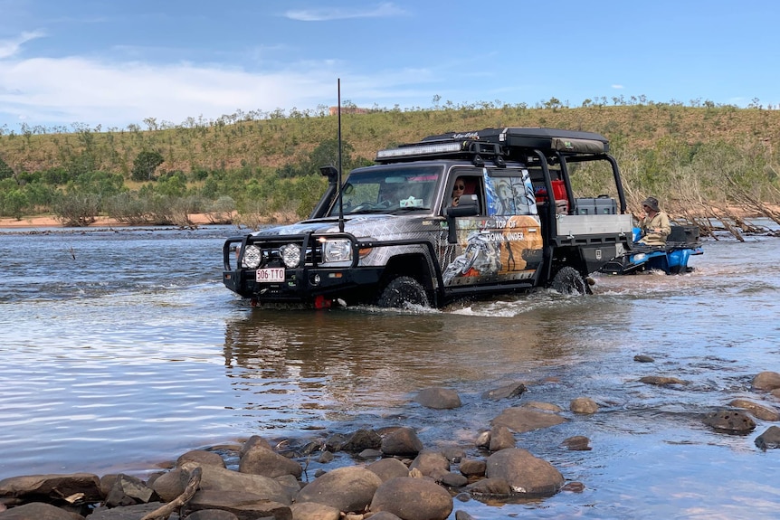 A four-wheel drive utility towing a man on a quad bike over a waterway.