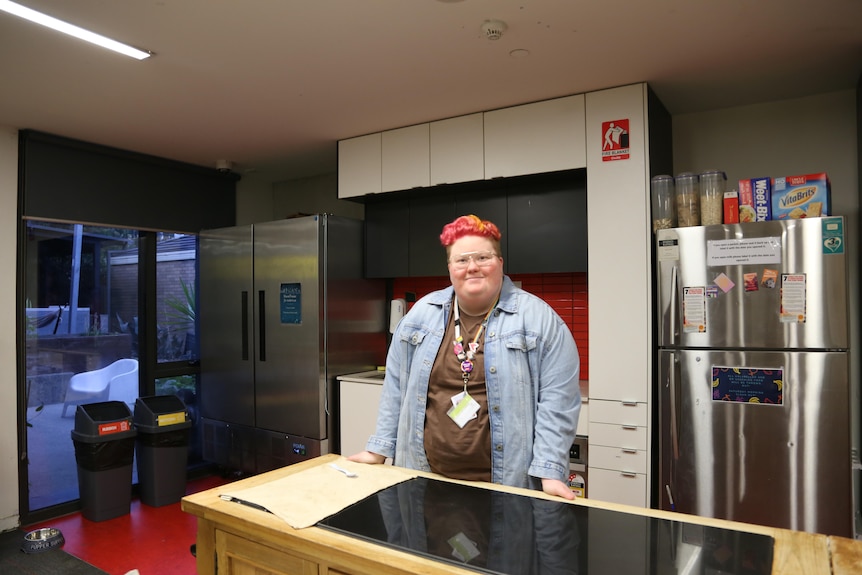 Non-binary young person with pink and yellow hair stands behind a kitchen counter in a communal living facility.